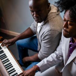 Two Black men sit at a piano. On the left, Benny's brow is furrowed. On the right, Gil looks down, but his face is peaceful.