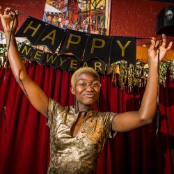 Satchel Williams, a young Black actor with cropped blonde hair, holds a a portion of a Happy New Year banner that reads "Happy" above their head.