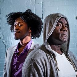 Two Black men stand one behind the other. In the foreground, Benny, played by Dwayne Alistair Thomas, looks directly at the camera with a more serious expression. Behind him, Gil, played by Garrick Vaughan, looks directly at the camera, his face more relaxed. Benny wears a grey hoodie. Gil wears a grey suit with a purple shirt.