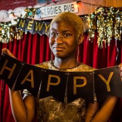 Satchel Williams, a young Black actor with cropped blonde hair, holds a a portion of a Happy New Year banner that reads "Happy" above their head.