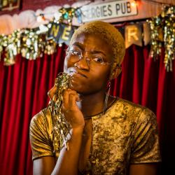 Satchel Williams, a young Black actor with cropped blonde hair is frowning, thinking, holding a piece of gold tinsel