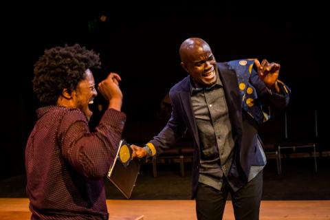 Two black men are smiling and dancing. On the left, Gil wears a burgundy dress shirt. On the right, Benny dances in a navy blazer with a blue and yellow wax print feature on the shoulder, worn over a grey dress shirt.
