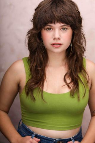 Anna Faye LIeberman's headshot. A young white person with wavy brown hair and bangs with brown eyes, wearing a green tank top and light makeup