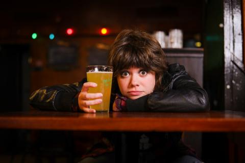 A young white woman rests her head on her arm on a bar, pint of beer in hand.